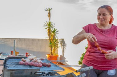 Kırmızı tişörtlü kızıl saçlı kadın evinin terasında barbekü yapıyor.