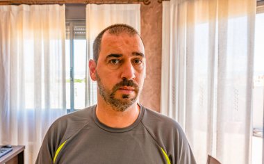 Evdeki oturma odasında poz veren kısa saçlı ve sakallı bir adam.