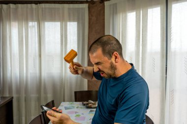 Sakallı ve kısa saçlı, lacivert gömlekli öfkeli bir adam cep telefonunu evdeki oturma odasına atıyor.