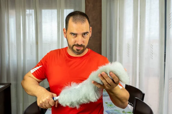 Man with a beard and short hair in a red shirt with a feather duster to clean the pole in the living room at home. Cleaning concept