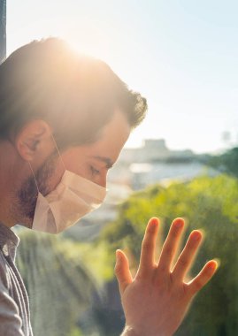 Coronavirus. Corona virüsünün hasta adamı pencereden bakıyor ve maske takıyor ve evdeki hastalıktan korunuyor. Karantinaya alın. Covid-19 koronavirüs salgını yüzünden çıkamadığı için üzgün. Pandemia. Ev.