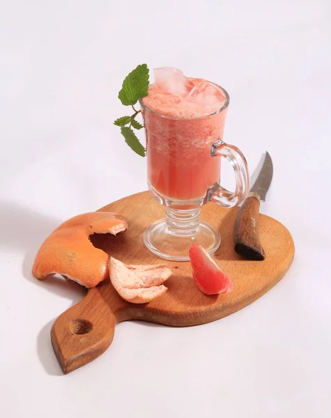 making grapefruit smoothie at home. Grapefruit smoothie in a glass cup and peel with a slot on a cutting board. Geypfruit leftovers and knife after making smoothie.