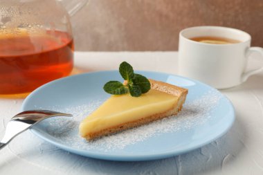 Limonlu tart dilimi, çaydanlık ve beyaz masada bir fincan çay.