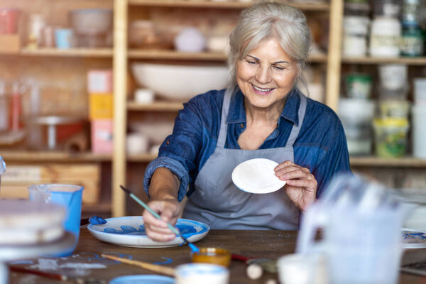 Зрелая ремесленница рисует тарелку из глины в художественной студии
