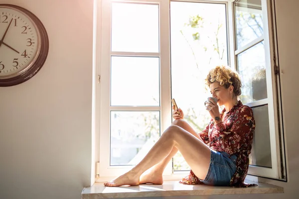 Young woman sitting on window sill and using smartphone