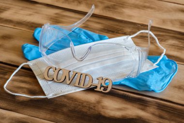 Beyaz tıbbi maske, mavi lastik eldivenler ve tahta bir yüzeyde güvenlik gözlükleri. Yazı kontrplak COVID-19 'dan yapılmış..