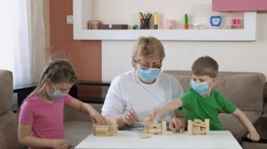 Büyükanne, tıbbi maskeli çocuklarla odada tahta bloklar ile oynuyor. COVID-19 salgını sırasında karantinada sosyal mesafeler ve kendini izole etme.
