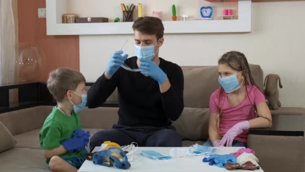 El hermano mayor muestra a los niños gafas protectoras y un respirador. Distanciamiento social y autoaislamiento en cuarentena durante la pandemia de COVID-19 . — Vídeos de Stock