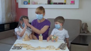 Tıbbi maskeli anne, oğul ve kız odalarında tahta yapboz oynuyorlar. Kuleleri yıkıyorlar. COVID-19 salgını sırasında karantinada sosyal mesafeler ve kendini izole etme.