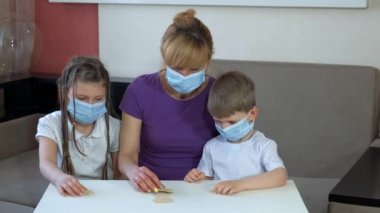 Tıbbi maskeli anne ve çocuklar odaya ahşap bulmacalar koydular. COVID-19 salgını sırasında karantinada sosyal mesafeler ve kendini izole etme.