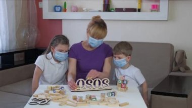 Tıbbi maskeli bir anne ve çocuklar içeride ahşap yapboz oynarlar. COVID-19 salgını sırasında karantinada sosyal mesafeler ve kendini izole etme.