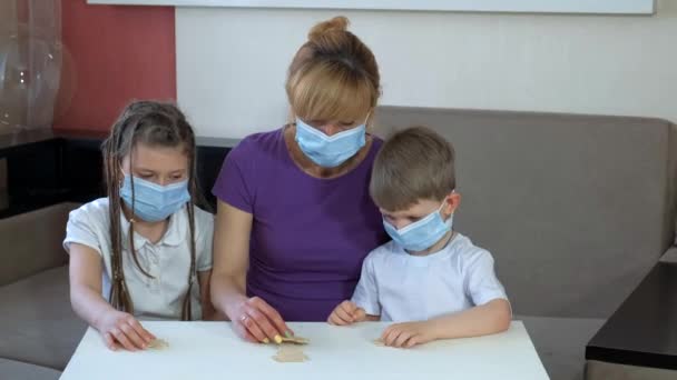 Madre e hijos en máscaras médicas armar rompecabezas de madera en la habitación. Distanciamiento social y autoaislamiento en cuarentena durante la pandemia de COVID-19 . — Vídeos de Stock