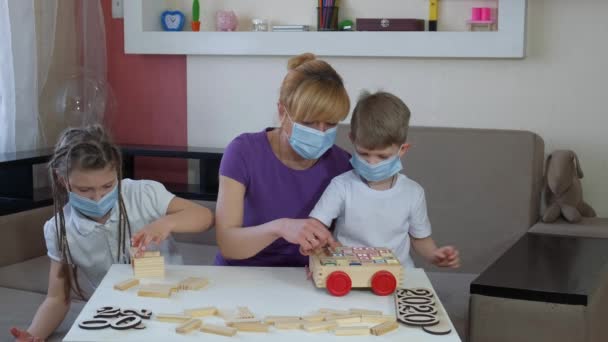 Madre, hijo e hija en máscaras médicas juegan rompecabezas de madera en la habitación. Distanciamiento social y autoaislamiento en cuarentena durante la pandemia de COVID-19 . — Vídeos de Stock