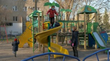 İki kız ve maskeli bir erkek çocuk oyun parkında kaydırağın üstünde kayıyorlar. Annem onlara yardım ediyor. Koronavirüs enfeksiyonlarına karşı koruma kavramı covid-19. 