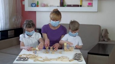 Tıbbi maskeli anne, oğul ve kız odalarında tahta yapboz oynuyorlar. COVID-19 salgını sırasında karantinada sosyal mesafeler ve kendini izole etme.