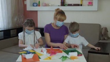 Tıp maskeli anne, oğul ve kız odadaki renkli kağıtlardan el işi yapıyorlar. COVID-19 salgını sırasında karantinada sosyal mesafeler ve kendini izole etme.