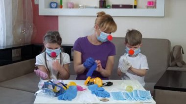 Tıbbî bandajlı anne ve çocuklar ve kırmızı palyaço burunlu çocuklar odada lastik eldiven giyerler. COVID-19 salgını sırasında karantinada sosyal mesafeler ve kendini izole etme.