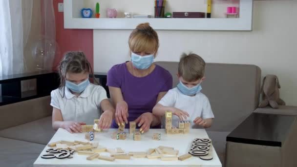 Mãe, filho e filha em máscaras médicas jogar quebra-cabeças de madeira na sala. Distanciamento social e autoisolamento em quarentena durante a pandemia de COVID-19 . — Vídeo de Stock