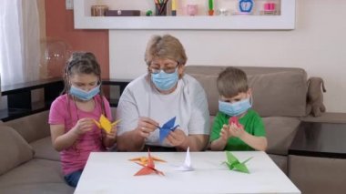 Tıbbi maskeli çocukların büyükannesi odada kağıt vinç yapıyor. COVID-19 salgını sırasında karantinada sosyal mesafeler ve kendini izole etme.