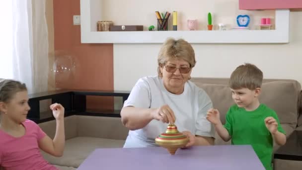 Büyükanne çocuklara odada dönen eski bir oyuncağın nasıl döndürüleceğini öğretiyor. COVID-19 salgını sırasında karantinada sosyal mesafeler ve kendini izole etme. — Stok video