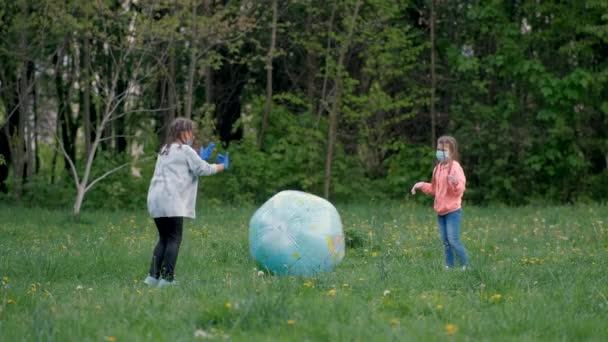 医療用マスクの2人の女の子が春に公園で大きなインフレータブルボールで遊ぶ。covid-19コロナウイルス感染からの保護の概念. — ストック動画