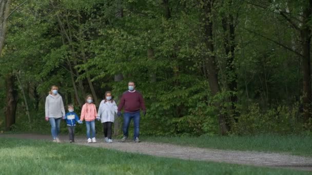 Wiosną w parku spaceruje rodzina w maskach medycznych. Ojciec i matka noszą dzieci w ramionach. Koncepcja ochrony przed zakażeniami covid-19 koronawirusami. — Wideo stockowe