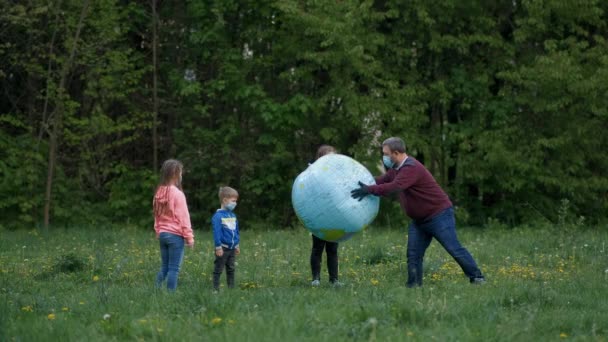 En far med barn i medicinska masker leker med en stor uppblåsbar boll i parken på våren. Begreppet skydd FROM covid-19 coronavirus infektioner. — Stockvideo