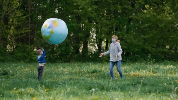 Een moeder en zoon met medische maskers spelen in het voorjaar met een groot opblaasbaar bal in het park. Het concept van bescherming van covid-19 coronavirus infecties. — Stockvideo