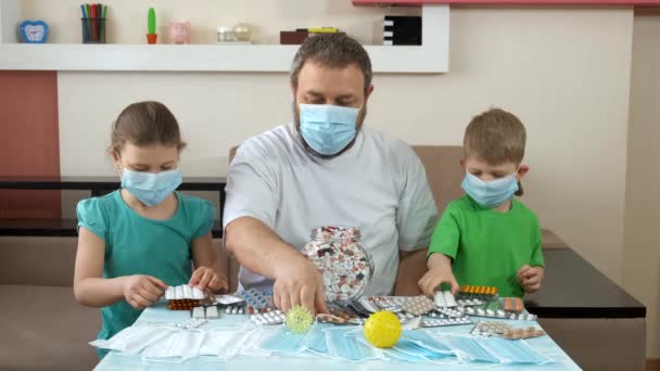 Un padre y sus hijos con máscaras médicas ponen pastillas sobre la mesa. Distanciamiento social y autoaislamiento en cuarentena durante la pandemia de COVID-19 . — Vídeos de Stock
