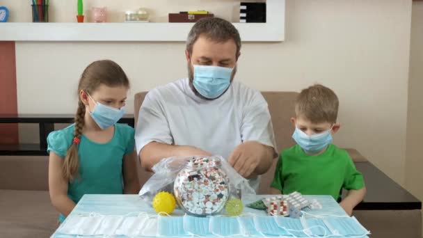 Een vader en kinderen met medische maskers leggen pillen op tafel. Sociale afstand en zelfisolatie in quarantaine tijdens de COVID-19 pandemie. — Stockvideo