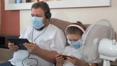 Baba ve kızı tıbbi maskeler ve kulaklıklar takıp oyun oynuyorlar. Yaz, sıcak, masa vantilatörünün yanında. COVID-19 salgını sırasında karantinada sosyal mesafeler ve kendini izole etme.