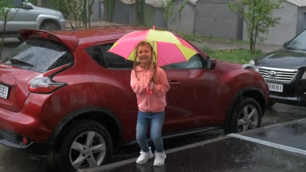 家の近くの駐車場の車の背景に雨の中で傘を持って陽気な女の子が喜んでジャンプします — ストック動画