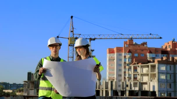 若いエンジニアの建築家男と女の子保護ヘルメットとベスト建設クレーンの背景に図面上のプロジェクトを見て — ストック動画