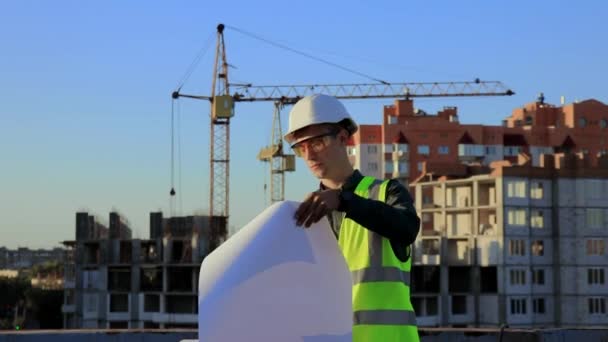一名戴着防护头盔和背心的年轻建筑工程师对照建筑用起重机的背景看了看图纸上的项目 — 图库视频影像