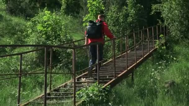 赤いジャケットと黒いバックパックを着た若い男が放棄された場所に移動します 彼は夏の日に森の古い錆びた階段を登り — ストック動画