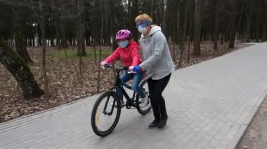 Annem kızına şehir parkında bisiklet sürmeyi öğretiyor. Koruyucu kasklar ve tıbbi maskeler takıyorlar. Covid-19 koronovirüs enfeksiyonundan korunma kavramı.