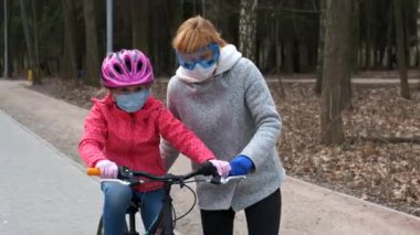 Annem kızına şehir parkında bisiklet sürmeyi öğretiyor. Koruyucu kasklar ve tıbbi maskeler takıyorlar. Covid-19 koronovirüs enfeksiyonundan korunma kavramı.