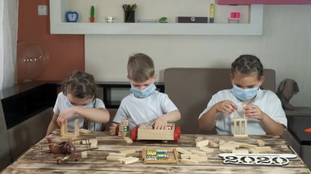 2人の女の子と医療用マスクの男の子が部屋に木製のおもちゃやパズルで遊んでいます Covid 19パンデミック時の隔離における社会的距離と自己隔離 — ストック動画