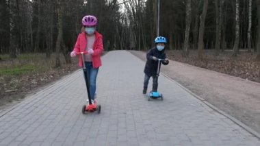 Küçük bir oğlan ve kız parkta maske takıp scooter 'a biniyorlar. Covid-19 koronavirüs enfeksiyonlarından korunma kavramı.