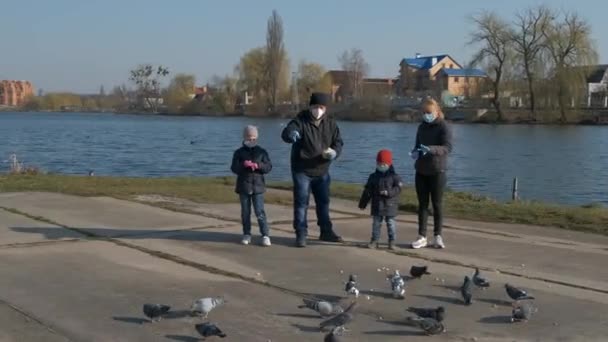 Mama Papa Sohn und Tochter in medizinischen Masken füttern Tauben in einem Park am See. Konzept der Unterstützung von Tieren mit COVID-19 Coronavirus.