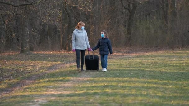 母と娘はスーツケース付きの医療用マスクで春の公園を歩きます コロナウイルス感染に対する保護の概念 Covid — ストック動画
