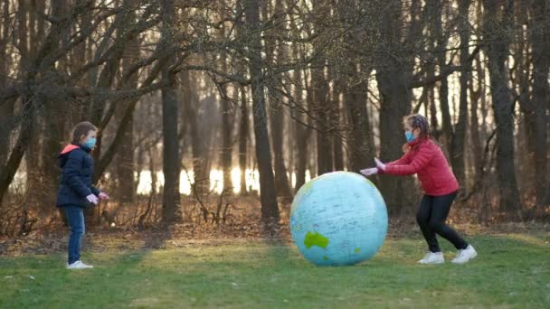 医療用マスクの2人の女の子は 大きなインフレータブルボール惑星地球と春の公園で再生されます コロナウイルス感染に対する保護の概念 Covid — ストック動画