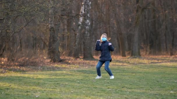 公園の春には小さな女の子が医療マスクで踊ります 犬は棒で走り — ストック動画