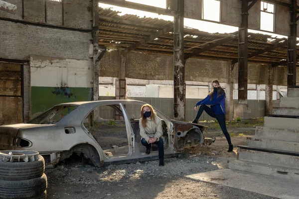 Siyah maskeli iki genç kız yıkılmış bir binanın içinde, bir arabanın kalıntılarının yanında. Kıyametin sonuçları, salgın, yıkım..