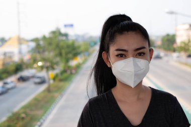 Hava yoluyla bulaşan solunum hastalıklarından korumak için N95 maskesi takan bir kadına yakından bakın. Grip vid-19 koronavirüs ebola Pm2.5 tozu ve yoldaki dumanlar arka planda patlamış.