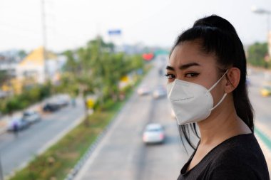 Hava yoluyla bulaşan solunum hastalıklarından korumak için N95 maskesi takan bir kadına yakından bakın. Grip vid-19 koronavirüs ebola Pm2.5 tozu ve yoldaki dumanlar arka planda patlamış.