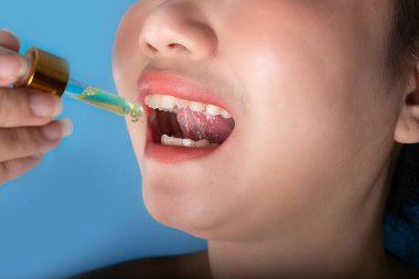 Beautiful Asea young woman holding a dropper pipette sublingual Cannabis oil at blue background, Sick or unwell young women using a drop on his mouth for medicine, health and beauty concept clipart