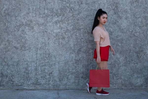Full length portrait of a beautiful young Asia woman holding shopping bags at concrete wall background, Women wear pink clothes and red skirt looking at the camera