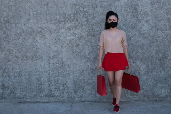 Full length portrait of a beautiful young Asia woman wearing a face mask with holding shopping bags at concrete wall background, Women wear pink clothes and red skirt looking at the camera