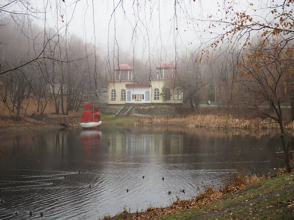 Gyönyörű őszi táj kilátással az erdőre a ködben és egy tó kacsákkal, egy gyönyörű régi épület és egy dekoratív hajó skarlátos vitorlákkal. Oroszország, Saratov - november, 2019 — Stock Fotó